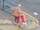 An elderly person with white hair is walking with the aid of a yellow cane. They are dressed in colorful clothing featuring a floral shirt and patterned pants. They are pulling a small cart filled with bundled newspapers. The person is also holding an empty Budweiser beer box. The scene is set on a paved area next to a blue metal fence with a yellow road marking curving alongside.