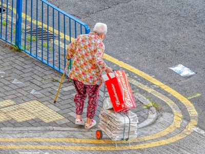 An elderly person with white hair is walking with the aid of a yellow cane. They are dressed in colorful clothing featuring a floral shirt and patterned pants. They are pulling a small cart filled with bundled newspapers. The person is also holding an empty Budweiser beer box. The scene is set on a paved area next to a blue metal fence with a yellow road marking curving alongside.