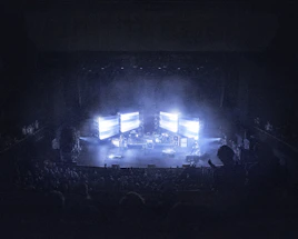 A concert stage is dimly lit, surrounded by an audience. Large screens display bright lights, creating an atmospheric effect. Musical instruments and equipment are set up on stage, contributing to a live performance setting.