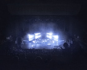 A concert stage is dimly lit, surrounded by an audience. Large screens display bright lights, creating an atmospheric effect. Musical instruments and equipment are set up on stage, contributing to a live performance setting.
