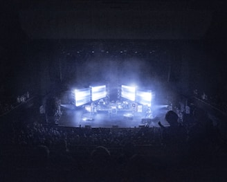 A concert stage is dimly lit, surrounded by an audience. Large screens display bright lights, creating an atmospheric effect. Musical instruments and equipment are set up on stage, contributing to a live performance setting.