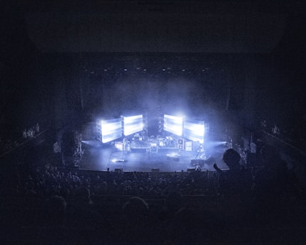 A concert stage is dimly lit, surrounded by an audience. Large screens display bright lights, creating an atmospheric effect. Musical instruments and equipment are set up on stage, contributing to a live performance setting.