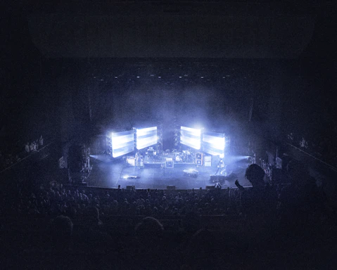 A concert stage is dimly lit, surrounded by an audience. Large screens display bright lights, creating an atmospheric effect. Musical instruments and equipment are set up on stage, contributing to a live performance setting.