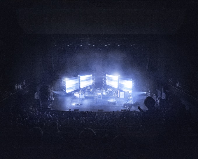A concert stage is dimly lit, surrounded by an audience. Large screens display bright lights, creating an atmospheric effect. Musical instruments and equipment are set up on stage, contributing to a live performance setting.