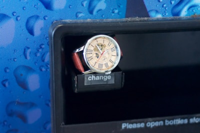 A wristwatch with a beige face and Roman numerals is resting on a slot labeled 'change', against a patterned blue background with a design resembling water droplets.