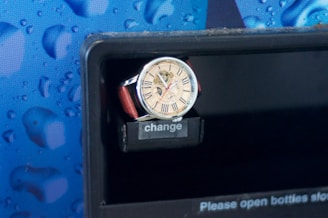 A wristwatch with a beige face and Roman numerals is resting on a slot labeled 'change', against a patterned blue background with a design resembling water droplets.