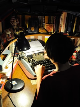 A person is seated at a wooden desk, typing on a vintage typewriter. The workspace is cluttered with books, framed photos, and small ornaments. A desk lamp casts a warm glow over the scene, highlighting the typewriter and the person's hands.