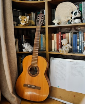 A wooden guitar is leaning on a bookshelf filled with an assortment of books and various stuffed animals. There's a woven hat and paper documents hanging down next to the guitar. The background features a curtain on the left side.
