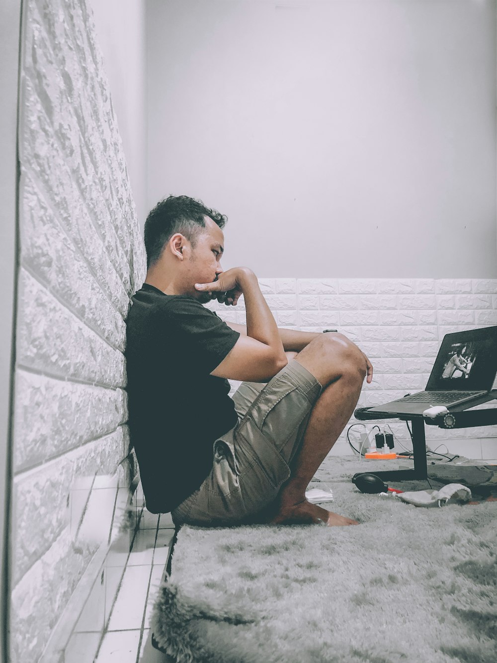 man wearing black t-shirt and brown cargo shorts sitting on floor watching movie on laptop with stand