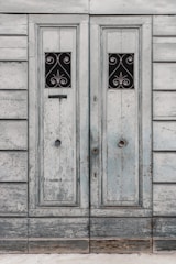 A pair of old, wooden doors with a weathered appearance, featuring ornate, wrought iron designs in small rectangular windows. The wood is faded, with a distressed texture, highlighting years of exposure.