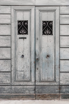 A pair of old, wooden doors with a weathered appearance, featuring ornate, wrought iron designs in small rectangular windows. The wood is faded, with a distressed texture, highlighting years of exposure.