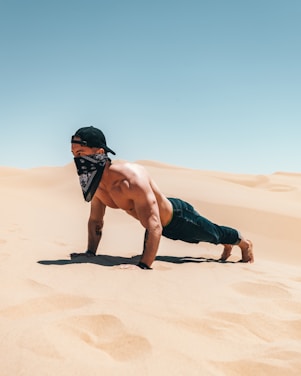 A shirtless man in jeans and a bandana performs a plank exercise in a vast desert landscape under a clear blue sky. He wears a black cap and is positioned on the sandy terrain, conveying a sense of strength and adventure.