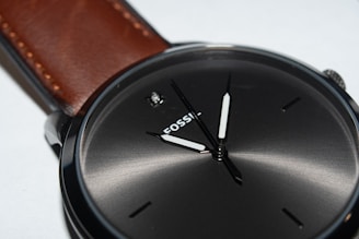 A close-up of a sleek wristwatch with a dark, metallic face and minimalistic hour markers. The watch has a simple, elegant design with a brown leather strap attached to a polished metal case.
