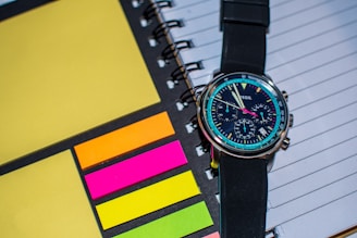 A wristwatch with a black strap is placed on top of a spiral-bound notebook. The notebook has lined pages and is accompanied by colorful sticky notes in shades of yellow, orange, pink, and green. The watch face is detailed with multiple dials and markings, showing modern and sleek design.