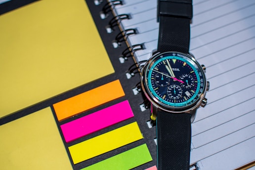 A wristwatch with a black strap is placed on top of a spiral-bound notebook. The notebook has lined pages and is accompanied by colorful sticky notes in shades of yellow, orange, pink, and green. The watch face is detailed with multiple dials and markings, showing modern and sleek design.