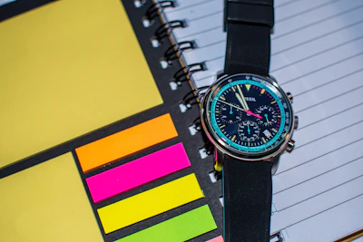 A sleek, modern watch with a black metal mesh strap lying next to a leather sketchbook.