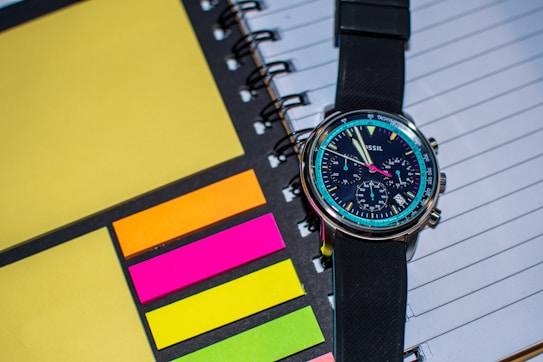 A wristwatch with a black strap is placed on top of a spiral-bound notebook. The notebook has lined pages and is accompanied by colorful sticky notes in shades of yellow, orange, pink, and green. The watch face is detailed with multiple dials and markings, showing modern and sleek design.