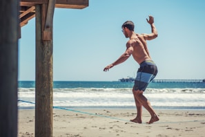 A man is balancing on a slackline at a beach. He is shirtless and wearing shorts, appearing focused as he maintains his balance. The ocean waves roll in the background, with a pier visible further down the shore. Wooden beams from a structure are partially visible to the left.