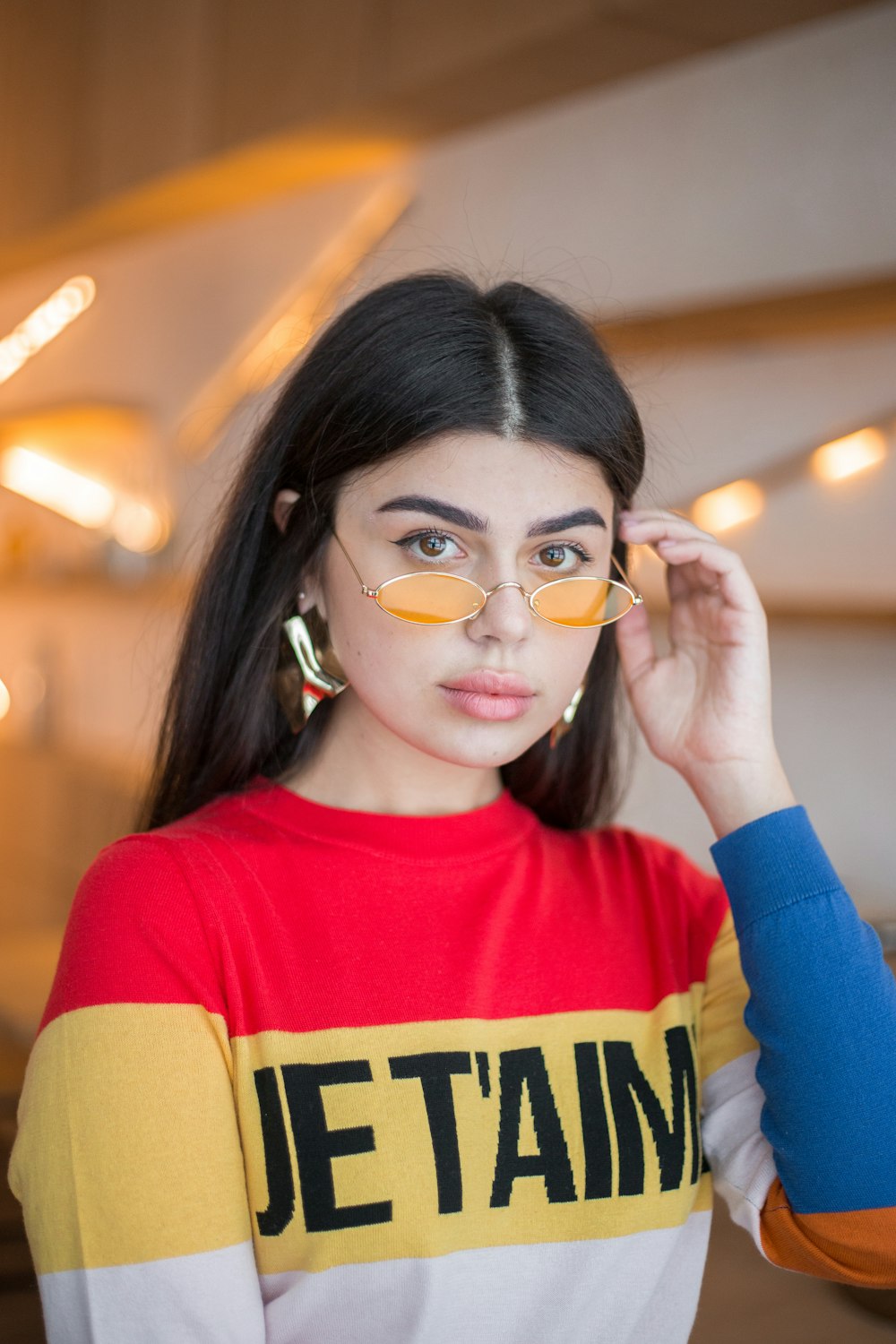 Femme portant une chemise à manches longues rouge, jaune et blanche tenant ses lunettes de soleil