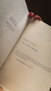 A hand with red nail polish is holding open a book, revealing a dedication and a page titled 'PART ONE: The Girl Who Cried Wolf.' Below is a quote from Ray Bradbury's 'Fahrenheit 451.'