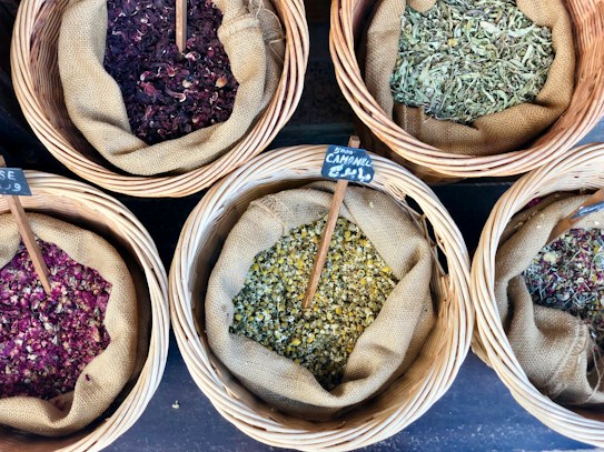 Several wicker baskets filled with various dried herbs and flowers, each with a small wooden sign labeling the contents. The herbs are displayed on a dark surface, and the baskets have a rustic appeal.