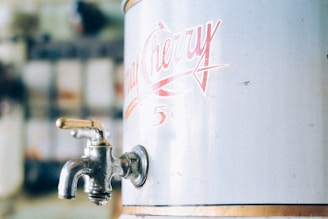 A close-up of a metal dispenser with a vintage style, featuring a brass tap on the left side and a retro logo in red lettering on the right side.