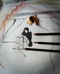 A pencil drawing on paper featuring an abstract silhouette of a person sitting and reading a book. Three pencils and a pencil shaving are placed on the sketch. The background includes soft, curved pencil strokes in varied colors such as gray and hints of brown and red, creating a layered texture.