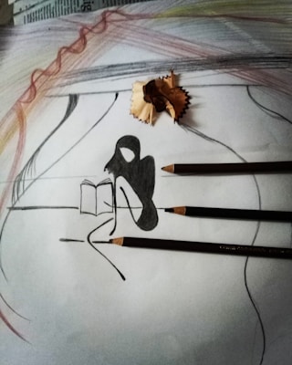 A pencil drawing on paper featuring an abstract silhouette of a person sitting and reading a book. Three pencils and a pencil shaving are placed on the sketch. The background includes soft, curved pencil strokes in varied colors such as gray and hints of brown and red, creating a layered texture.