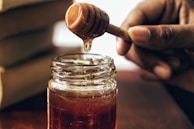 A jar filled with golden honey sits on a wooden surface. A person's hand holds a honey dipper above the jar as honey drips down. The setting appears warm and inviting, with natural lighting highlighting the rich color of the honey.