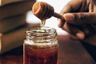 A jar filled with golden honey sits on a wooden surface. A person's hand holds a honey dipper above the jar as honey drips down. The setting appears warm and inviting, with natural lighting highlighting the rich color of the honey.