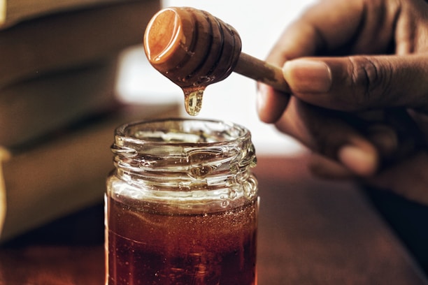 A jar filled with golden honey sits on a wooden surface. A person's hand holds a honey dipper above the jar as honey drips down. The setting appears warm and inviting, with natural lighting highlighting the rich color of the honey.