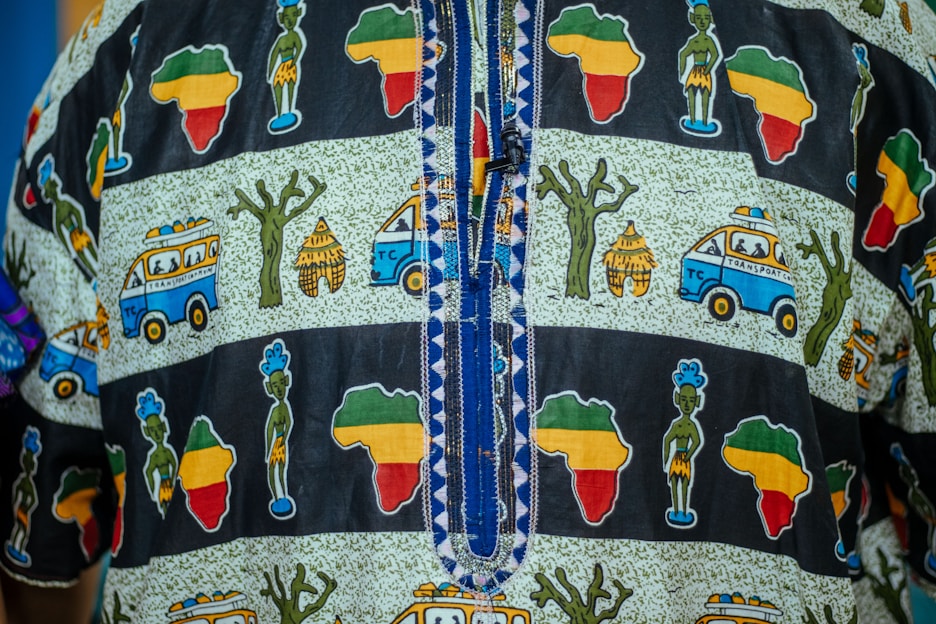 Colorful fabric with a pattern of African continent shapes in green, yellow, and red, alongside illustrations of people, trees, huts, and vehicles labeled as transportation. The fabric features geometric decorative stitching in the center.