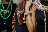 A group of individuals is adorned with colorful, intricately designed beaded necklaces and bracelets. The beads are strung in various vibrant colors, including green, yellow, pink, and blue, creating a striking contrast against the dark clothing they wear. The necklaces and bracelets have different patterns and sizes, showcasing a rich variety in design.