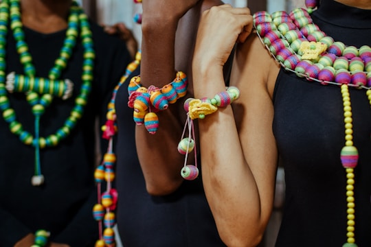 A group of individuals is adorned with colorful, intricately designed beaded necklaces and bracelets. The beads are strung in various vibrant colors, including green, yellow, pink, and blue, creating a striking contrast against the dark clothing they wear. The necklaces and bracelets have different patterns and sizes, showcasing a rich variety in design.