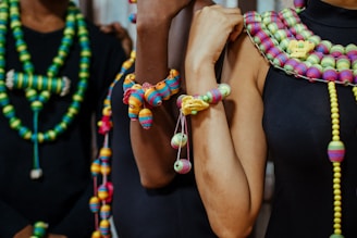 A group of individuals is adorned with colorful, intricately designed beaded necklaces and bracelets. The beads are strung in various vibrant colors, including green, yellow, pink, and blue, creating a striking contrast against the dark clothing they wear. The necklaces and bracelets have different patterns and sizes, showcasing a rich variety in design.