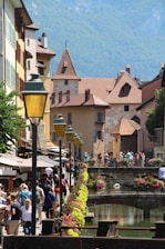 A picturesque European street scene featuring charming, historic buildings with red-tiled roofs and stone facades. A small canal lined with colorful flowers and decorative lamp posts runs along the pathway. People are strolling, enjoying a sunny day, creating a lively and vibrant atmosphere.