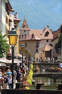 A picturesque European street scene featuring charming, historic buildings with red-tiled roofs and stone facades. A small canal lined with colorful flowers and decorative lamp posts runs along the pathway. People are strolling, enjoying a sunny day, creating a lively and vibrant atmosphere.
