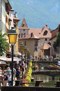 A picturesque European street scene featuring charming, historic buildings with red-tiled roofs and stone facades. A small canal lined with colorful flowers and decorative lamp posts runs along the pathway. People are strolling, enjoying a sunny day, creating a lively and vibrant atmosphere.