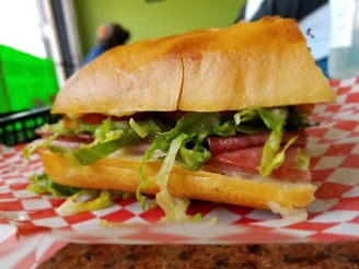 A close-up of a deli sandwich filled with fresh lettuce, slices of meat, and served on a crusty bun. The sandwich is placed on a checkered red and white paper, possibly on a wooden table. The background is slightly blurred, with hints of green and other muted colors.