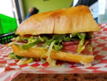 A close-up of a deli sandwich filled with fresh lettuce, slices of meat, and served on a crusty bun. The sandwich is placed on a checkered red and white paper, possibly on a wooden table. The background is slightly blurred, with hints of green and other muted colors.