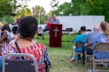 An outdoor gathering with people seated on chairs and a person speaking at a podium with a cross symbol. The background shows trees and a white truck in a park setting. The attendees appear to be attentively listening.
