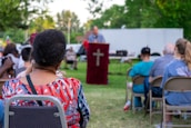 An outdoor gathering with people seated on chairs and a person speaking at a podium with a cross symbol. The background shows trees and a white truck in a park setting. The attendees appear to be attentively listening.