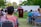 An outdoor gathering with people seated on chairs and a person speaking at a podium with a cross symbol. The background shows trees and a white truck in a park setting. The attendees appear to be attentively listening.