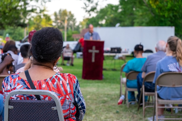An outdoor gathering with people seated on chairs and a person speaking at a podium with a cross symbol. The background shows trees and a white truck in a park setting. The attendees appear to be attentively listening.