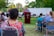 An outdoor gathering with people seated on chairs and a person speaking at a podium with a cross symbol. The background shows trees and a white truck in a park setting. The attendees appear to be attentively listening.