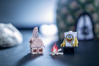 Two Lego minifigures resembling cartoon characters are seated around a small campfire. The background is blurred and includes a rock and a cave-like structure. The scene is set on a dark surface with a soft focus, implying an outdoor setting.