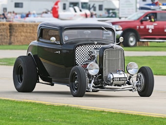A black vintage hot rod car with a checked grille and large, exposed headlights is parked on a paved road. The car features distinct, wide tires and a polished body, reflecting nearby surroundings. Behind the vehicle, there is a backdrop of grass, a red fire truck, and some people in the distance near temporary structures.