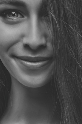 A close-up black and white portrait focusing on a person's lower face with a gentle smile and visible strands of hair. The image emphasizes the textures and expressions of the facial features, particularly the lips and part of the nose.