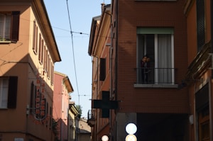 A narrow street flanked by buildings with warm-colored facades. An 'Enoteca' sign is visible on one of the buildings, indicating a wine shop. There is a person standing on a balcony looking out toward the street. The street appears calm, with no visible activity or traffic.