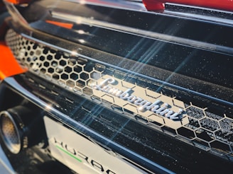 A close-up of a car featuring a honeycomb-patterned grille and the metallic logo of a luxury car brand prominently displayed. The image captures the play of light across the shiny surface, highlighting the premium finish and design elements.